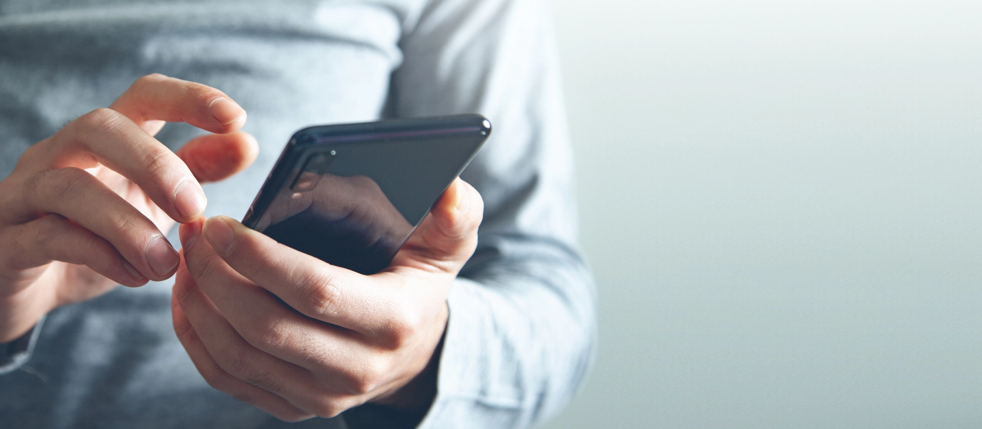 Nahaufnahme einer Person, die ein Smartphone in der Hand hält und den Touchscreen mit einer Hand bedient. Die Person trägt ein graues Langarmshirt, und der Hintergrund ist unscharf und hell gehalten