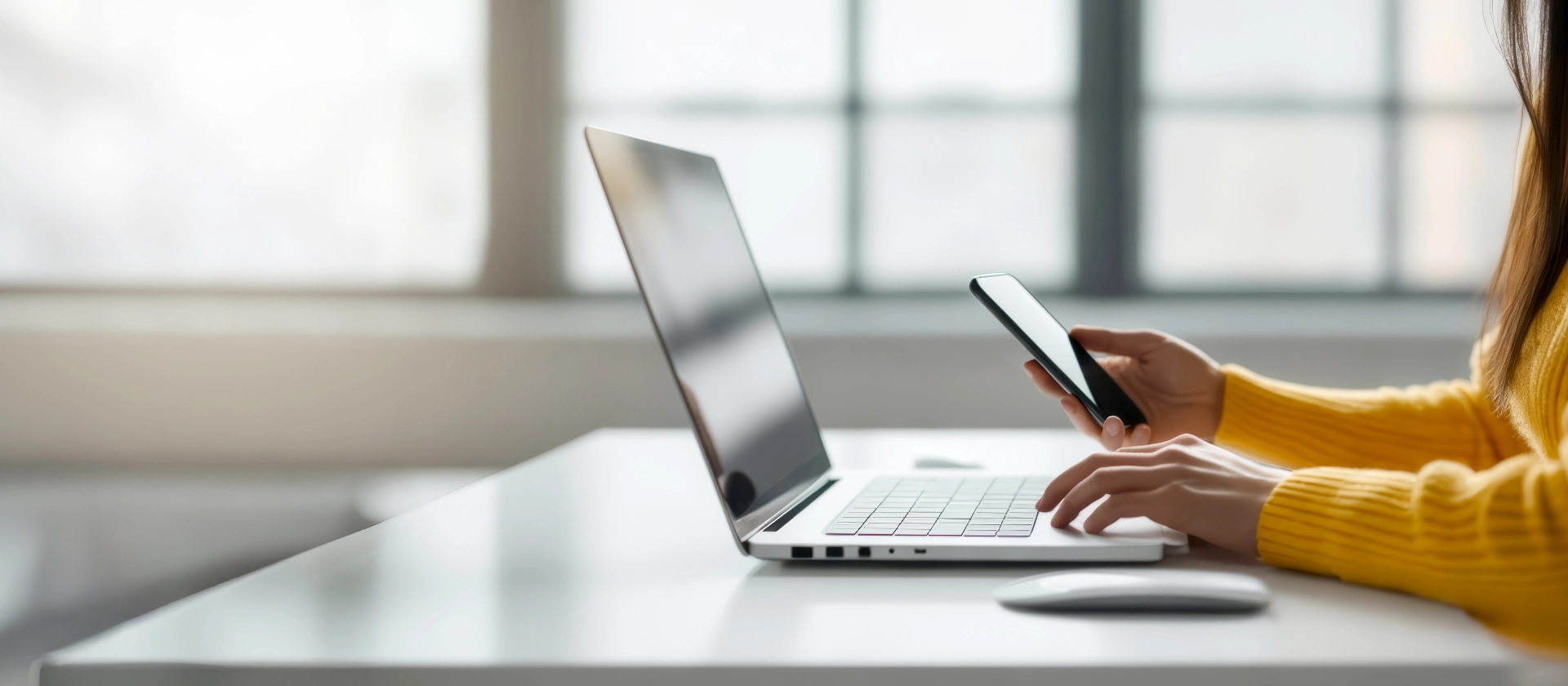 Eine Frau in einem gelben Pullover sitzt an einem weißen Schreibtisch und arbeitet an einem Laptop, während sie ein Smartphone in der Hand hält. Im Hintergrund ist ein großes Fenster mit weichem Tageslicht zu sehen.
