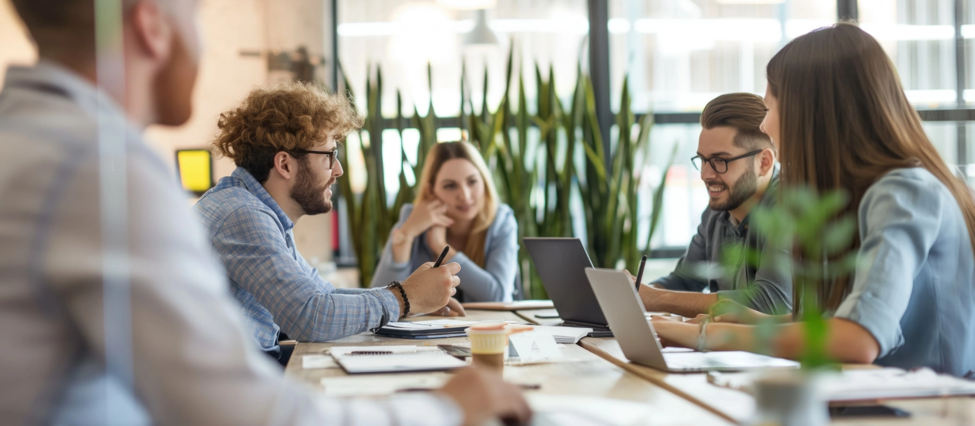Junge Erwachsene in einer Besprechung im modernen Büro. Kollegen sitzen um den Konferenztisch mit Laptops und besprechen die Projektstrategie.